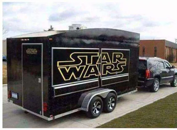 A black enclosed trailer with large gold 'STAR WARS' logo on its side is hitched to a black SUV parked on a paved surface…
