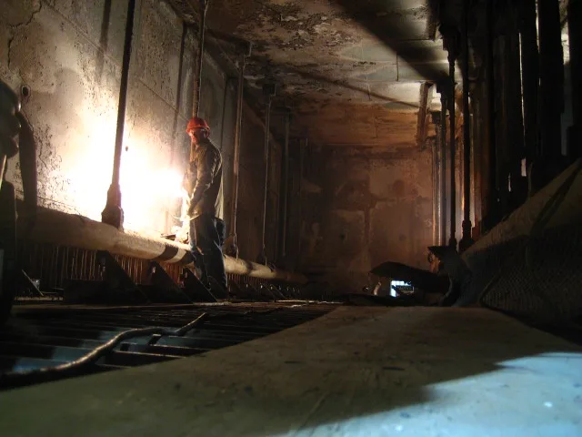 A low-angle shot of a worker in a hard hat welding in a dim, industrial tunnel, with bright light from the welding torch…