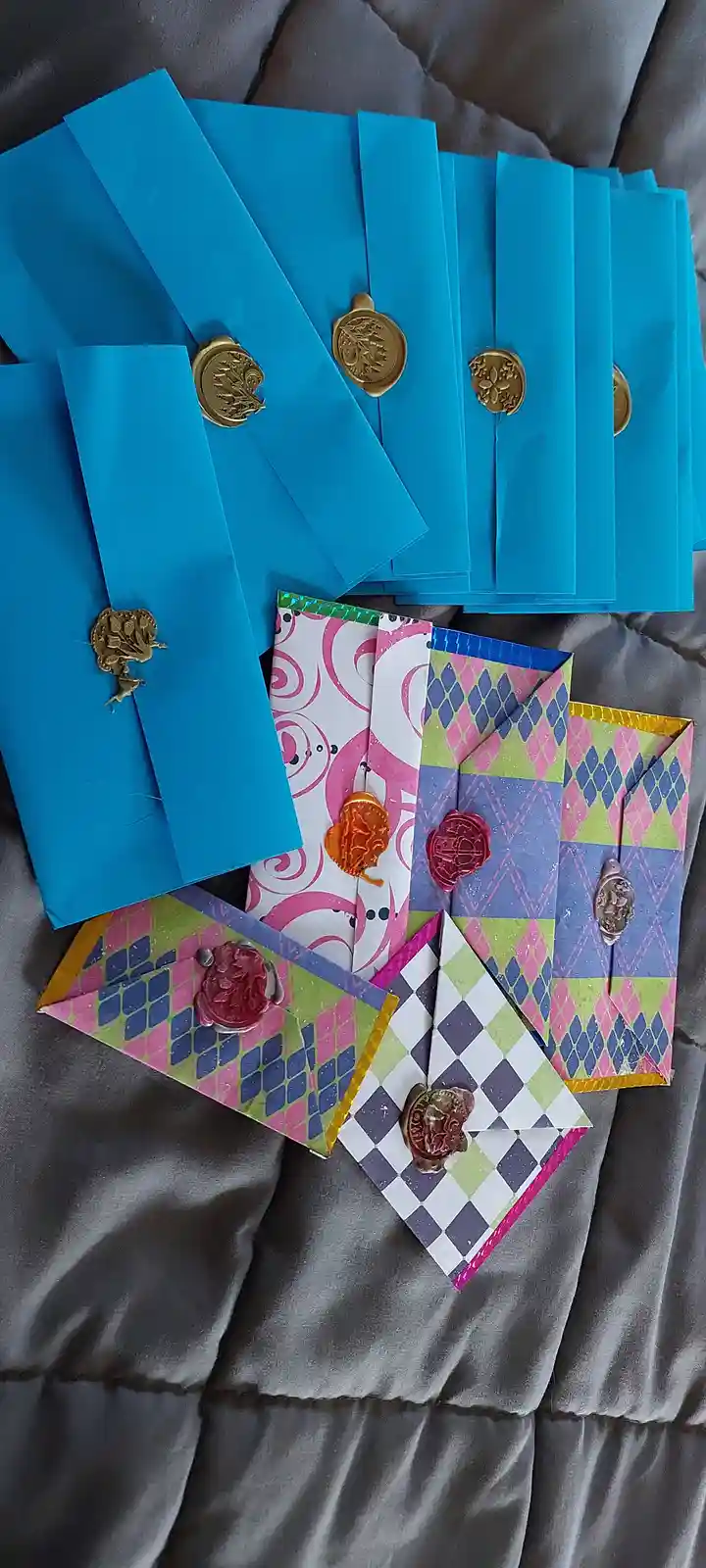 An overhead shot displays multiple handmade envelopes on a grey fabric surface. The top group consists of plain blue…