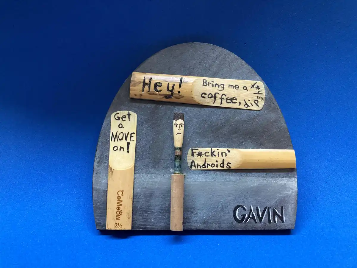 A handmade diorama on a dark gray curved board against a blue background, featuring small wooden pieces with handwritten…