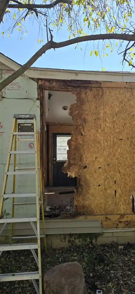 An exterior view of a house under renovation shows a large section of the wall sheetrock removed, revealing the wooden…