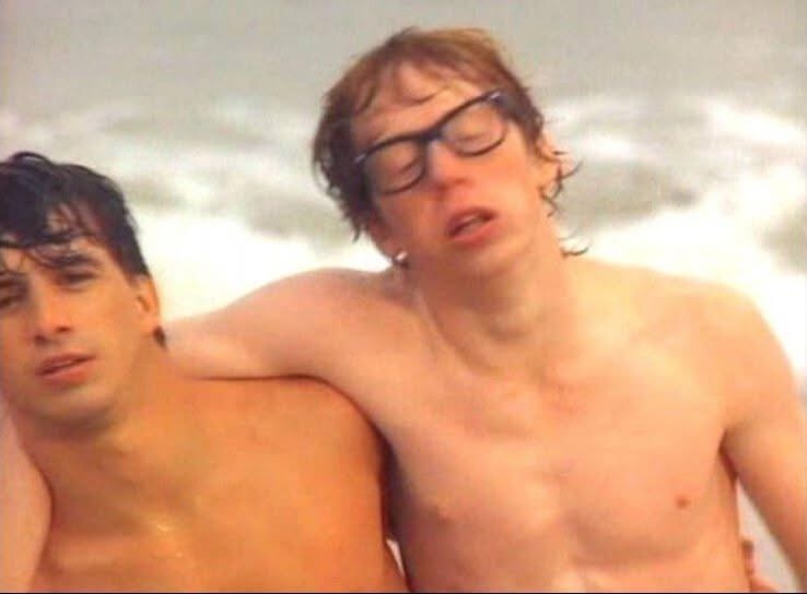Two shirtless men are in a blurry, grainy photo, likely from a beach or pool. The man on the right, wearing thick-rimmed…