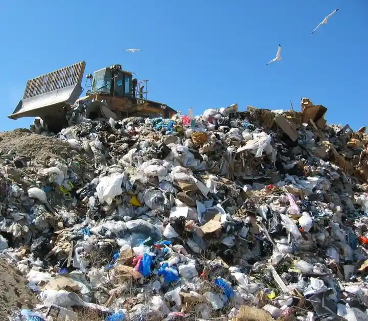 A large yellow bulldozer sits atop a massive, chaotic pile of trash under a clear blue sky with seagulls flying overhead…