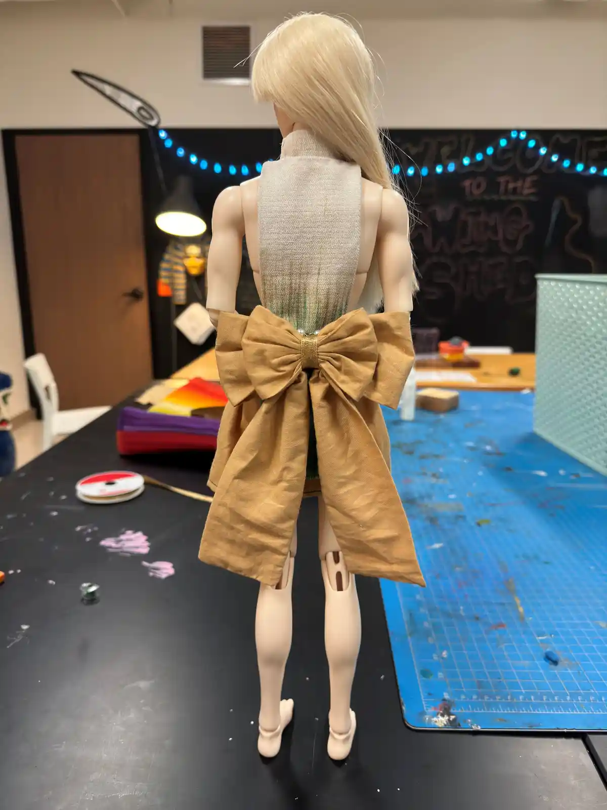 A doll with blonde hair stands with its back to the camera, wearing a beige fabric top and a large tan bow tied around its…