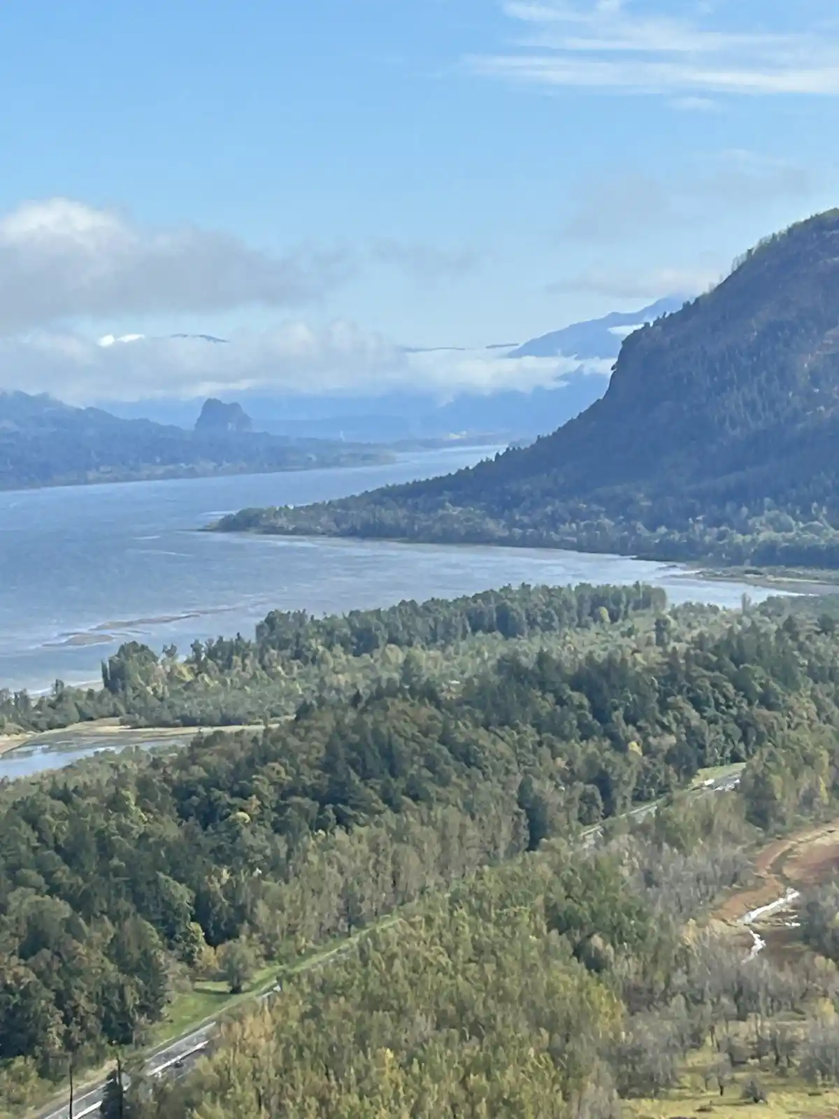 A scenic aerial view of a winding road through dense green forests, leading to a large body of water bordered by mountains…