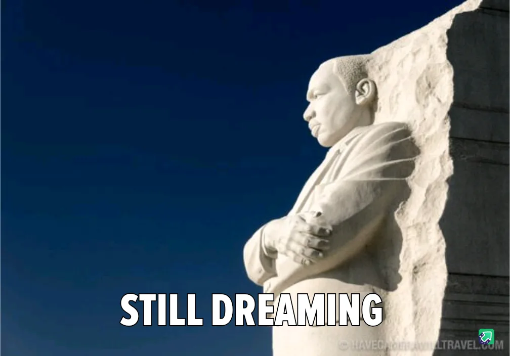 A stone statue of Martin Luther King Jr. is shown in profile against a clear blue sky, with his arms crossed and head…