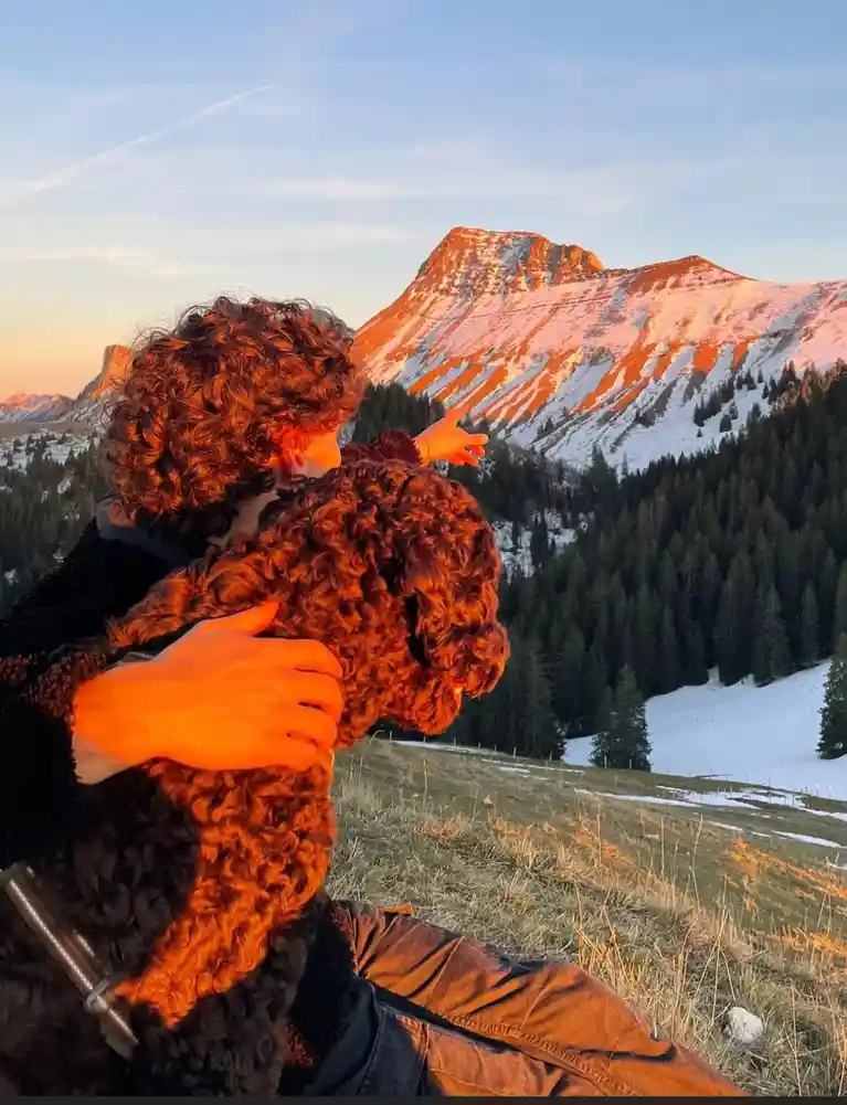A person with curly brown hair is sitting on a grassy hillside, hugging a large curly-haired dog. Both are bathed in the…