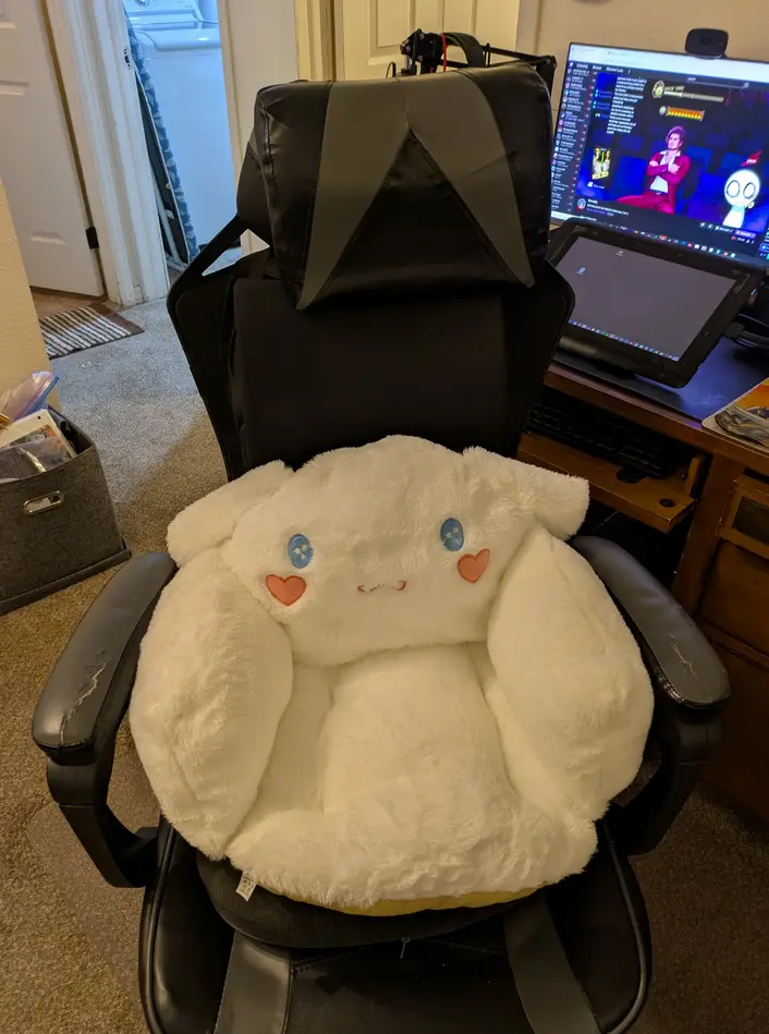 A black gaming chair has a large, fluffy white plush pillow shaped like a bunny with blue eyes and pink heart-shaped cheeks…