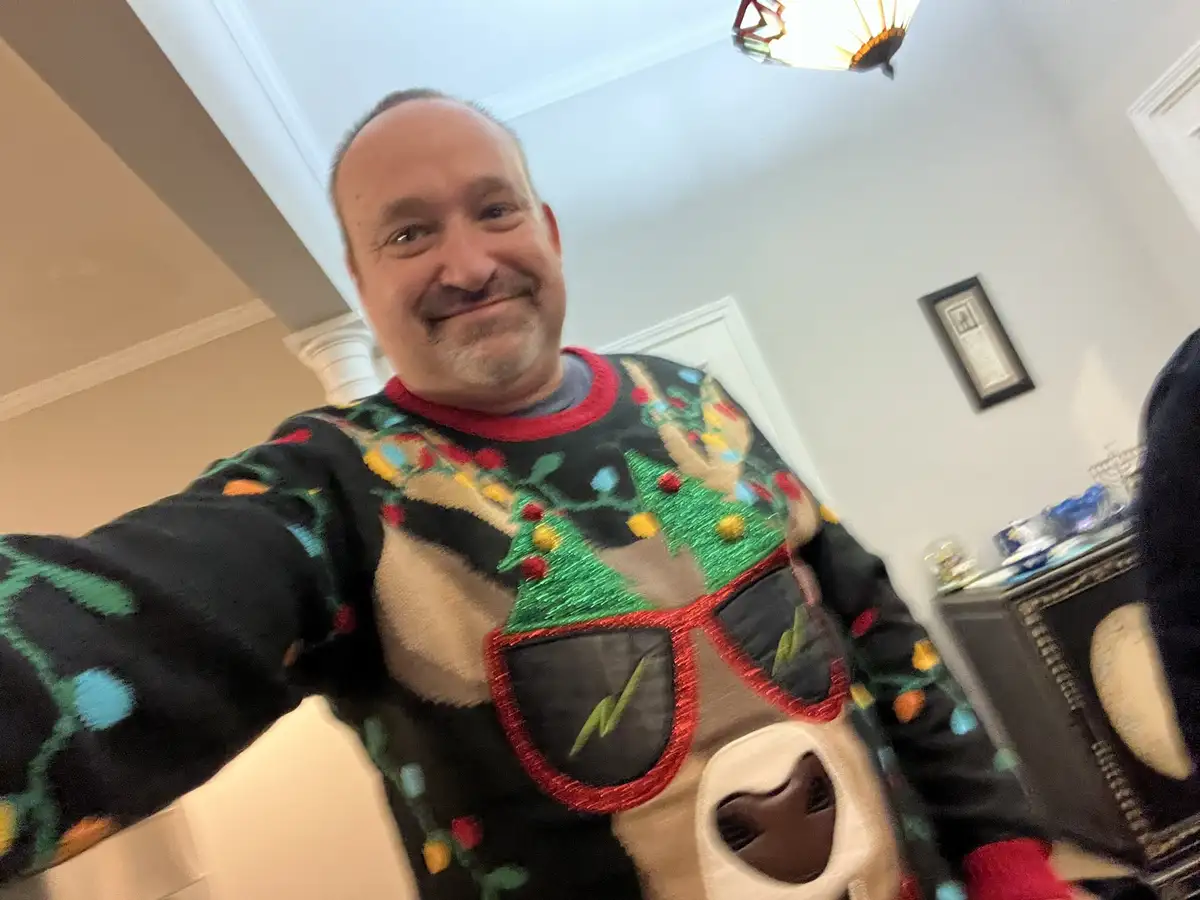 A man takes a selfie indoors wearing a very festive, overly decorated Christmas sweater featuring green trees, colorful…