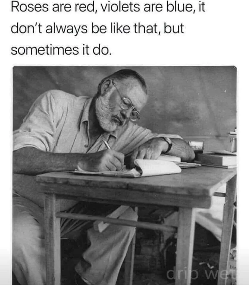 A black-and-white photo shows an older man with glasses and a beard, leaning over a wooden desk and writing in a notebook…