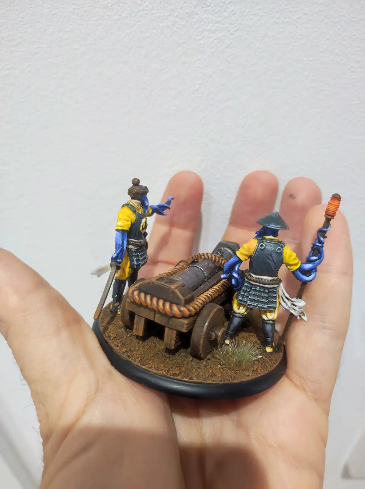 A hand holds a miniature diorama featuring two blue-skinned, yellow-and-black armored figures resembling samurai, one…