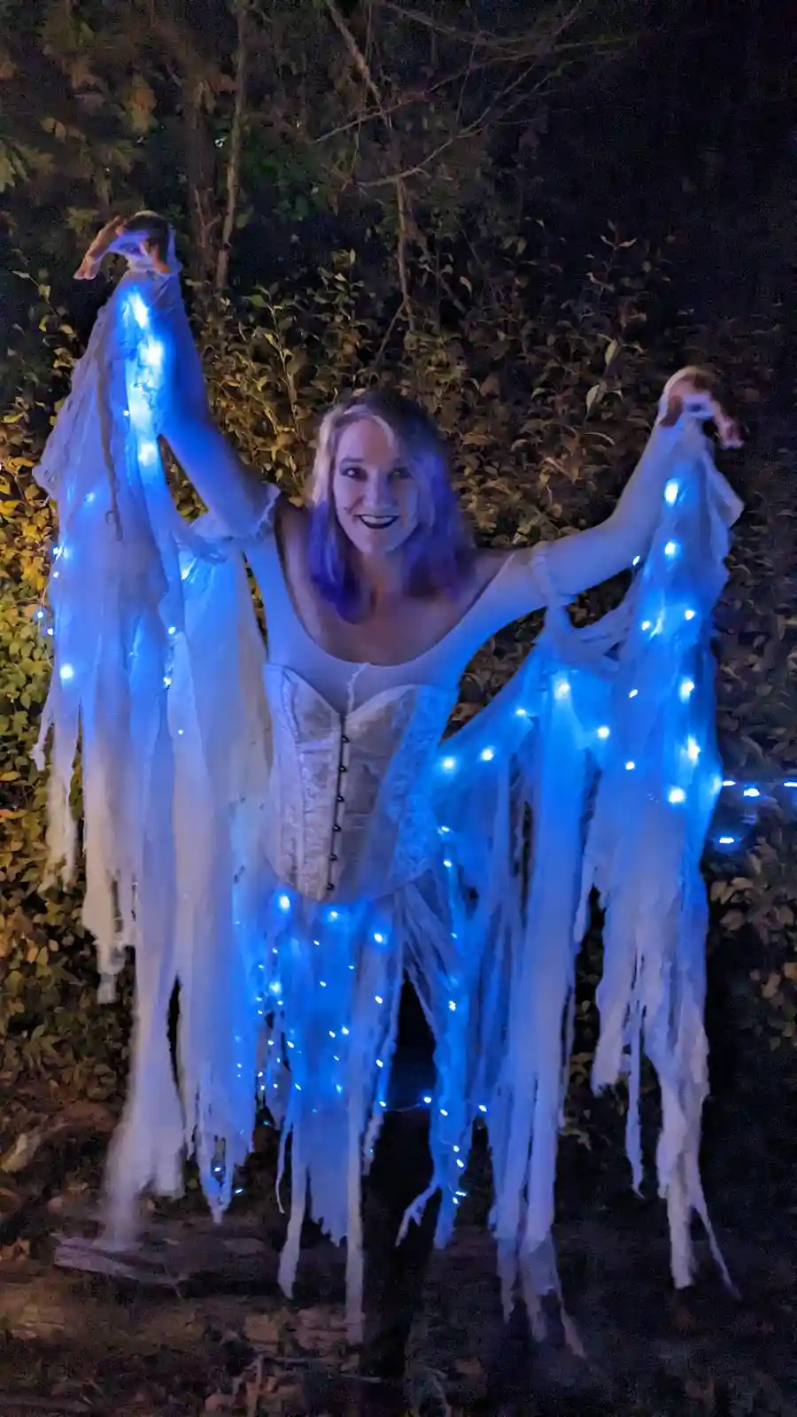 A person in a white corset and tattered white fabric costume stands outdoors at night, holding up the fabric which is strung…