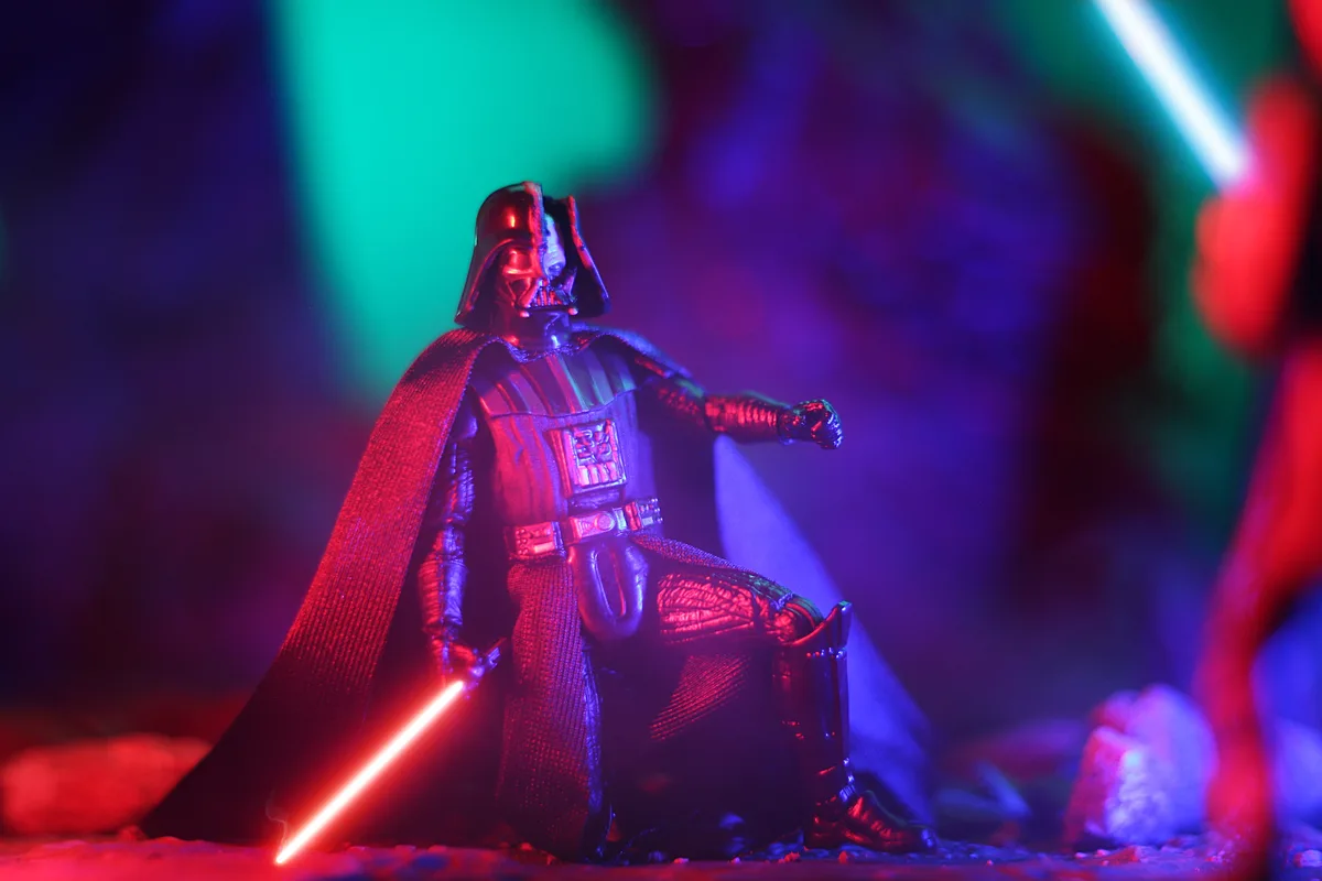 A Darth Vader action figure sits on a throne-like chair, holding a red lightsaber, under dramatic red and blue lighting…
