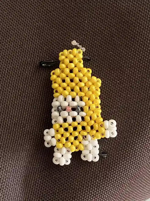 A handmade bead art figure, primarily yellow with white accents for limbs and face, resembling a cartoon character, is…