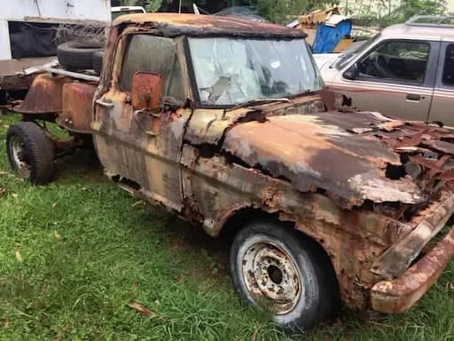 A heavily rusted, dilapidated pickup truck with missing panels and a broken windshield sits abandoned on grass. The overlaid…