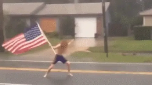A shirtless man with long hair is shown in six sequential frames running down a wet street while holding an American flag…