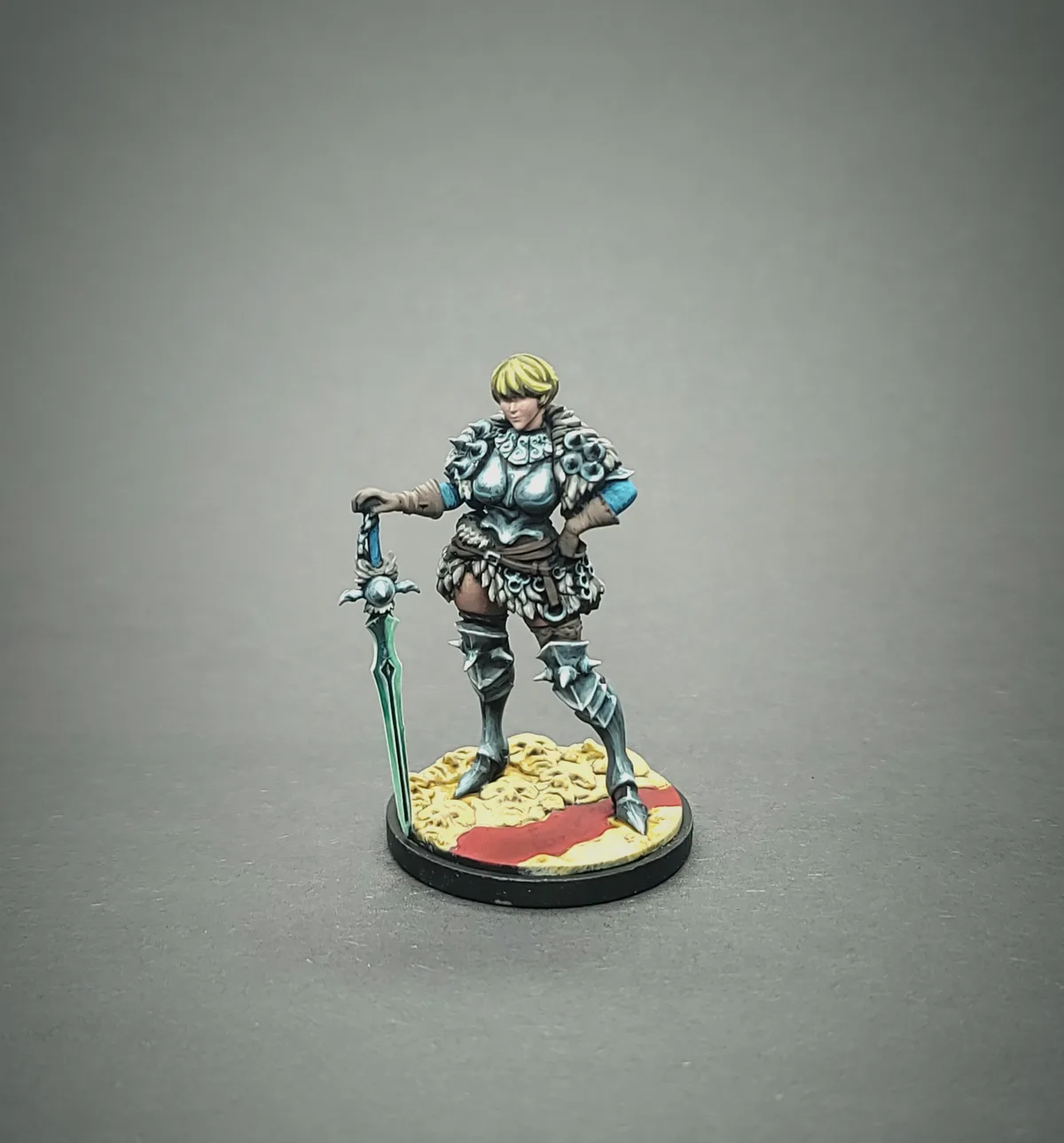 A painted miniature of a female warrior in ornate silver armor with blonde hair, standing on a circular base with a red and…