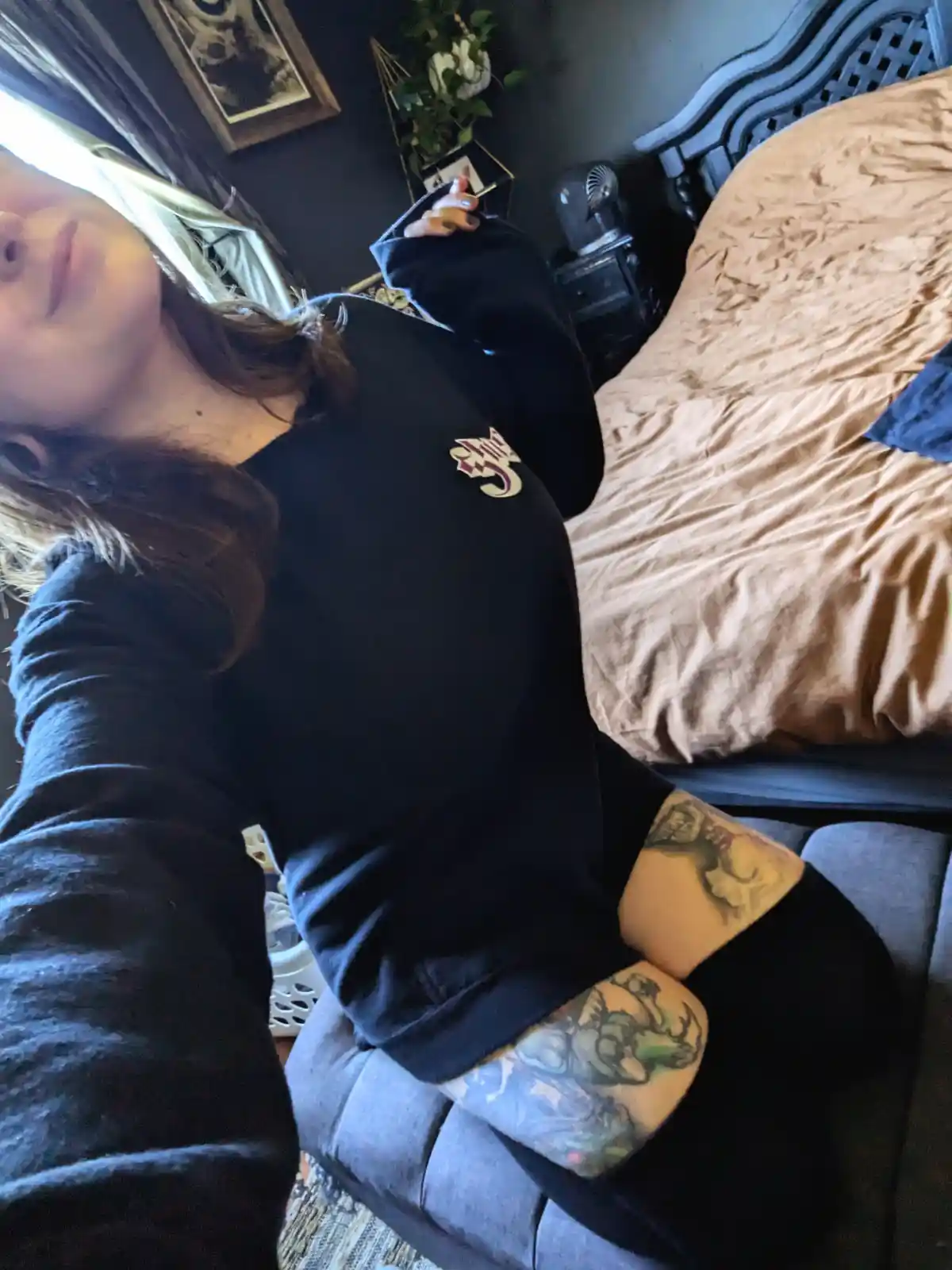 A person with visible tattoos on their arms is lying on a couch in a bedroom, wearing a black sweatshirt and looking up at…