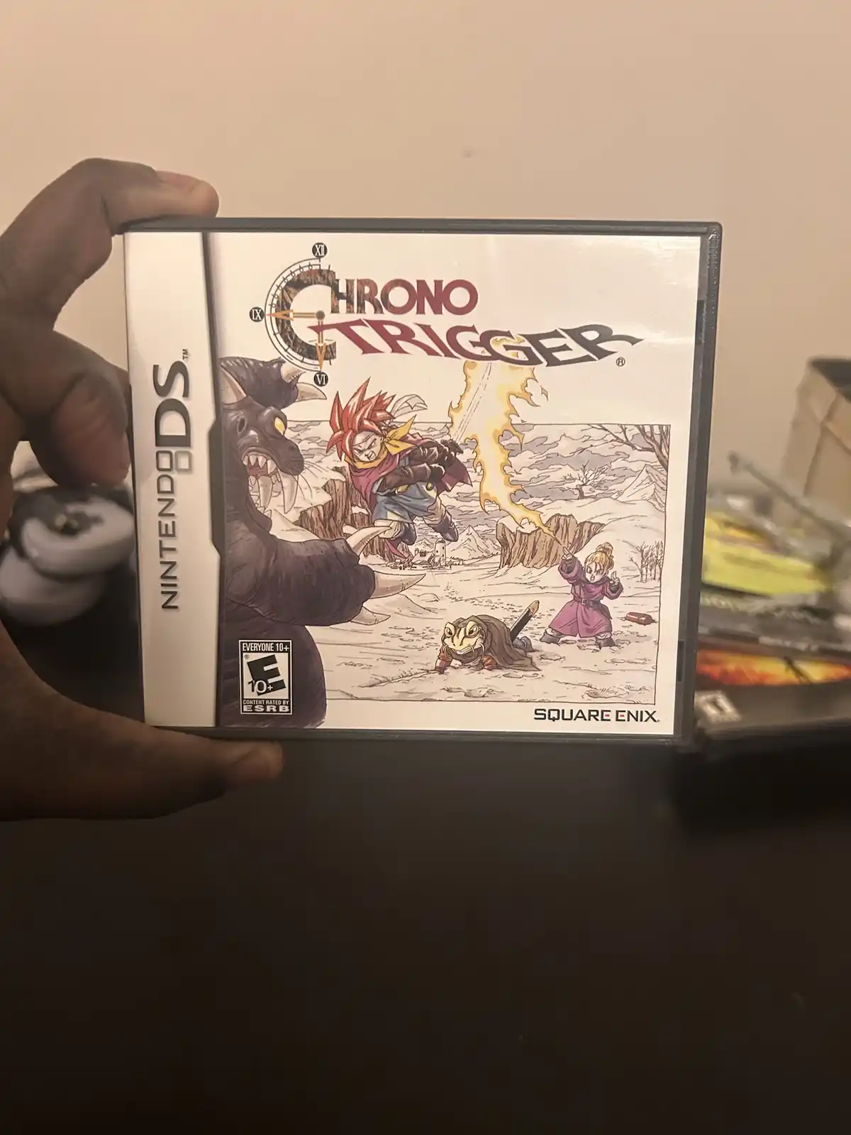A hand holds a Nintendo DS game case for Chrono Trigger, showing its cover art with characters in a snowy battle scene. The…