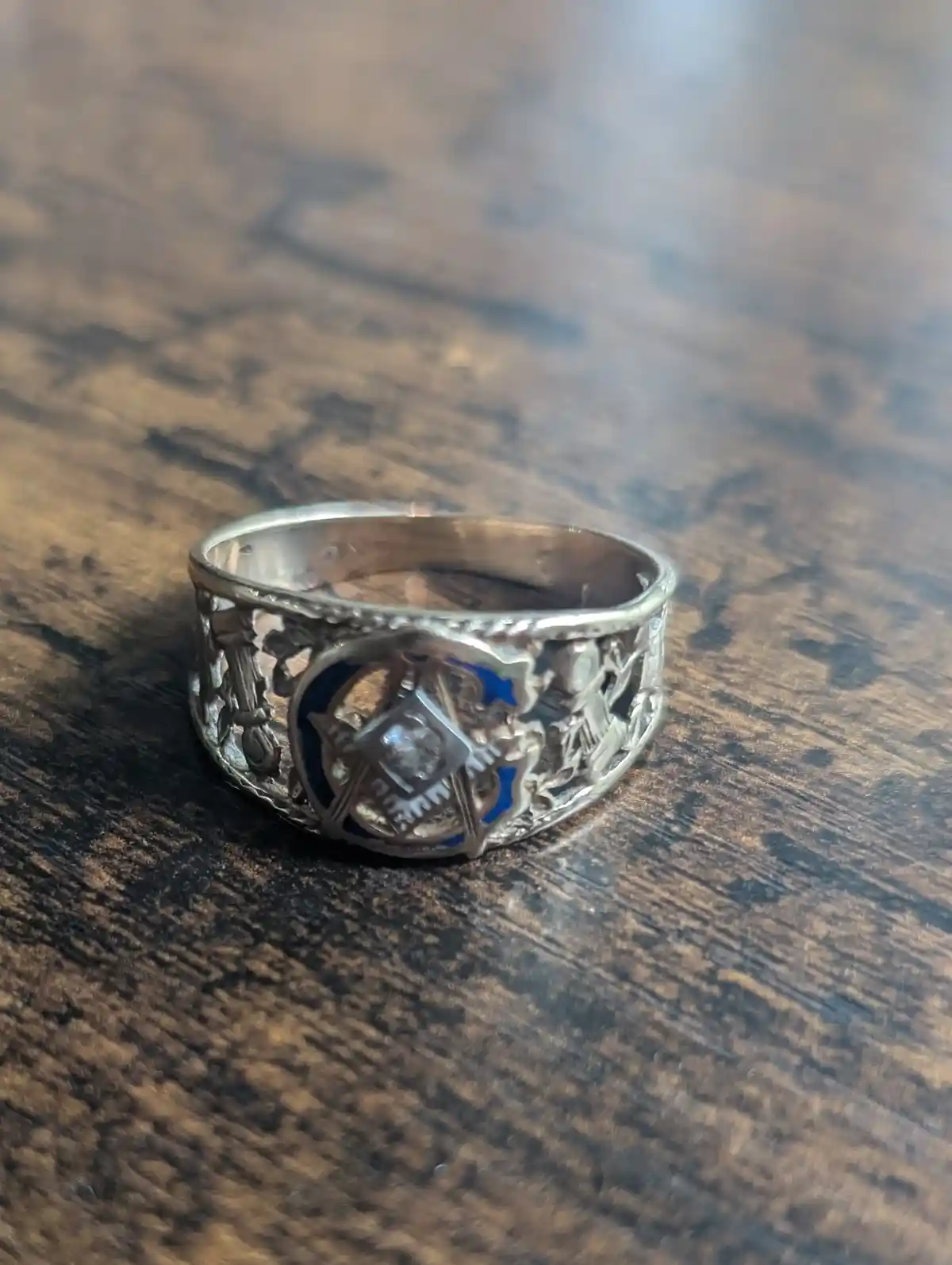A close-up photograph shows a silver Masonic ring with intricate filigree and a central blue enamel square and compasses…