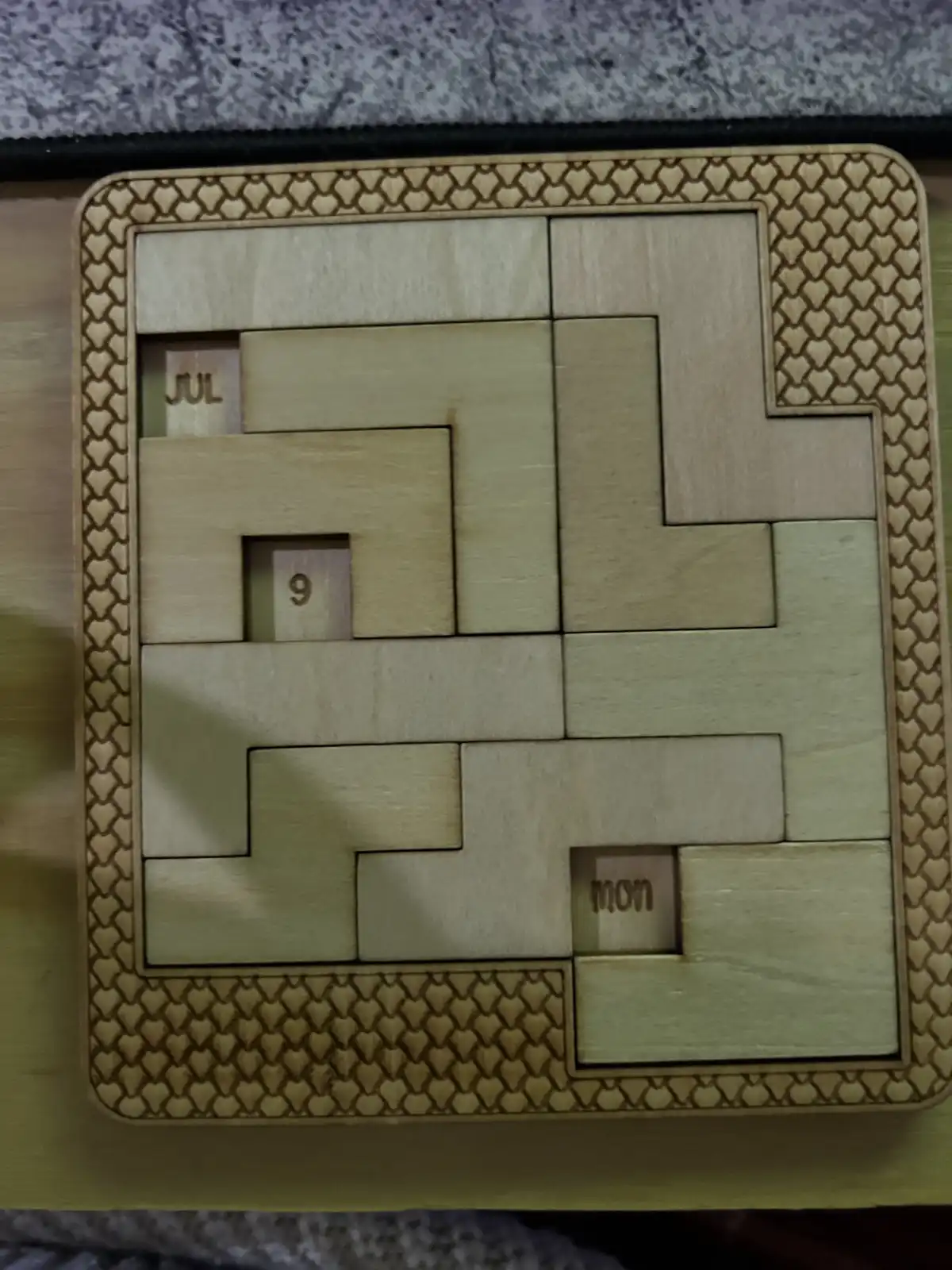 A wooden puzzle with interlocking pieces forms a grid, with three small squares showing the text 'JUL', '9', and 'MON' in…