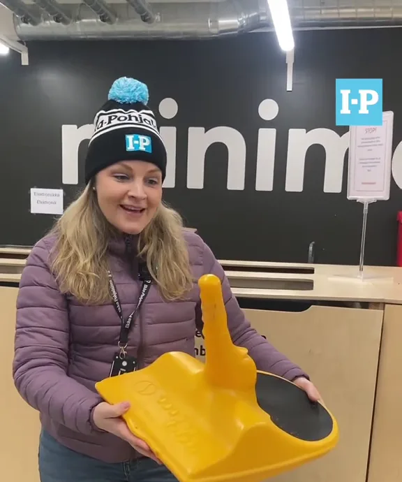 A woman wearing a black beanie with a blue pom-pom and an 'I-P' logo smiles while holding a large yellow plastic object…