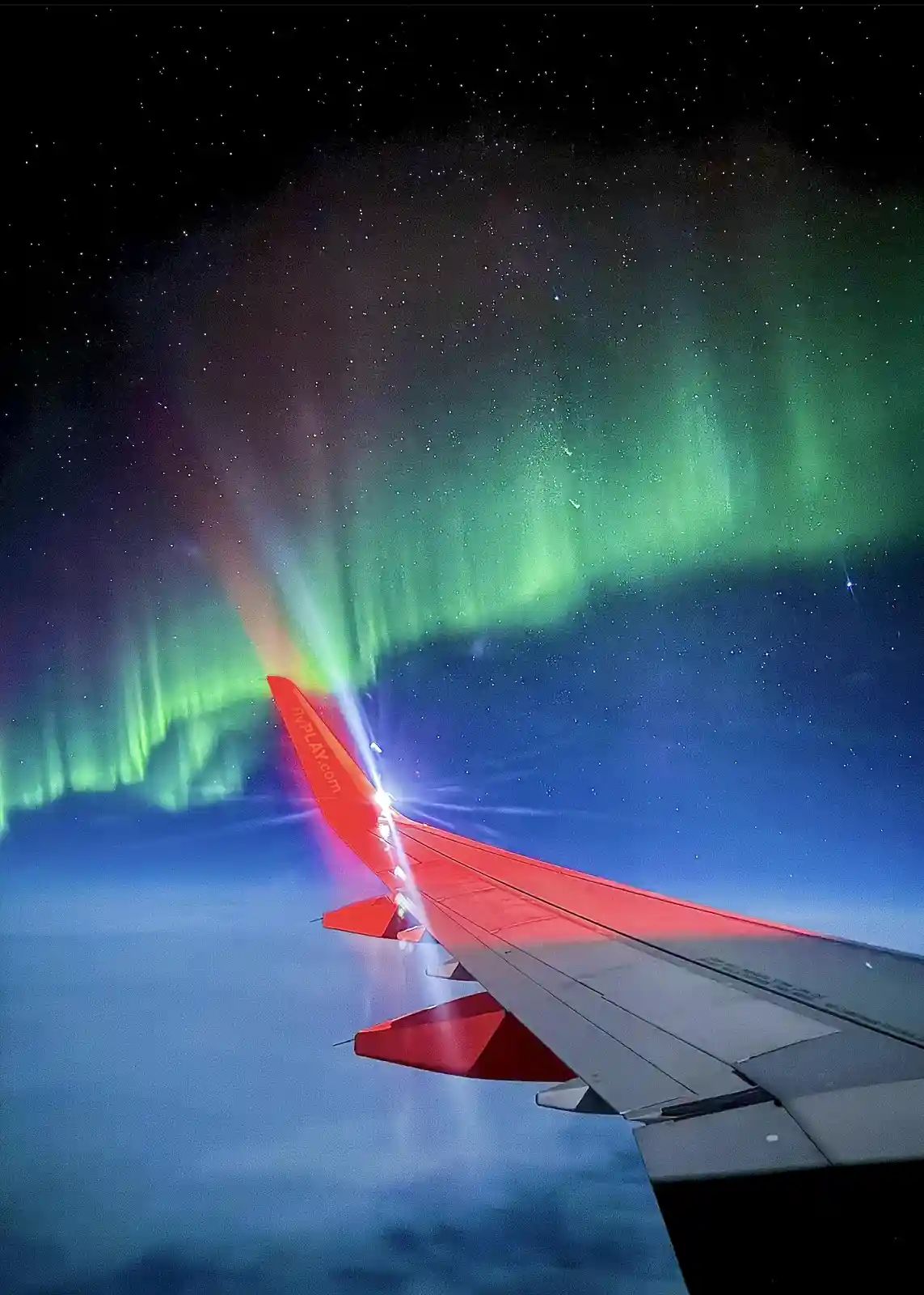 A view from an airplane window shows the red-tipped wing of a FlyPlay.com aircraft flying above clouds under a starry night…