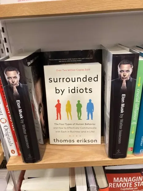 A bookshelf displays a book titled 'surrounded by idiots' by Thomas Erikson, flanked on both sides by copies of 'Elon Musk'…
