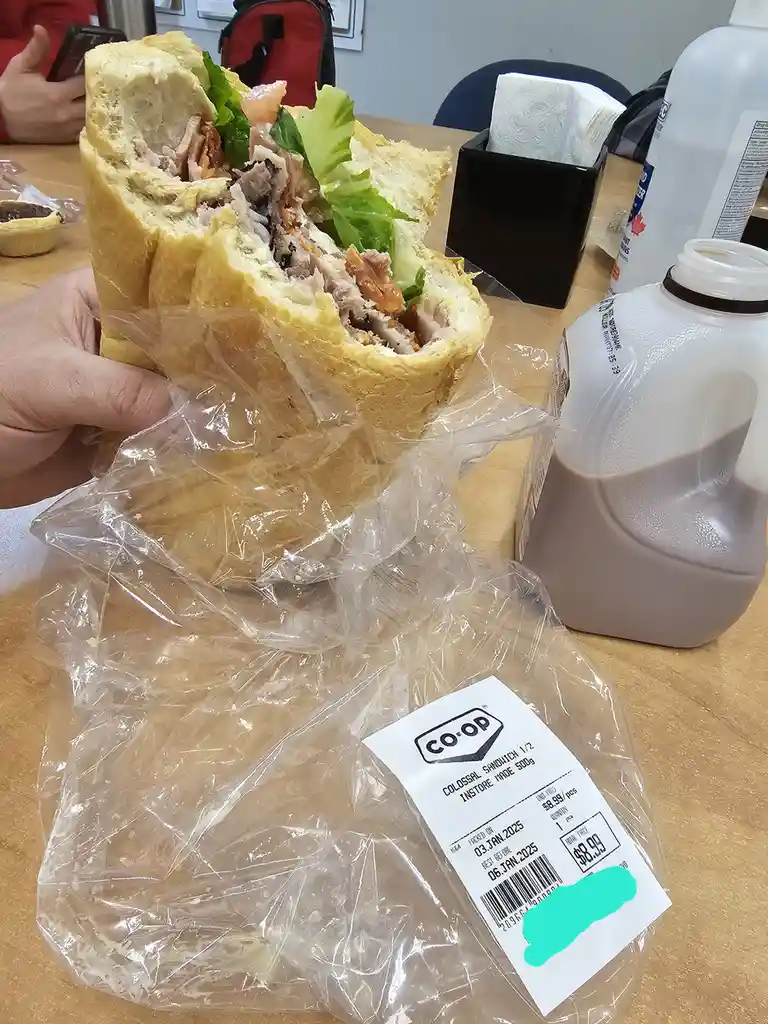 A hand holds a large, half-eaten sandwich wrapped in plastic, with a Co-op store label showing it's a 'Colossal Sandwich…