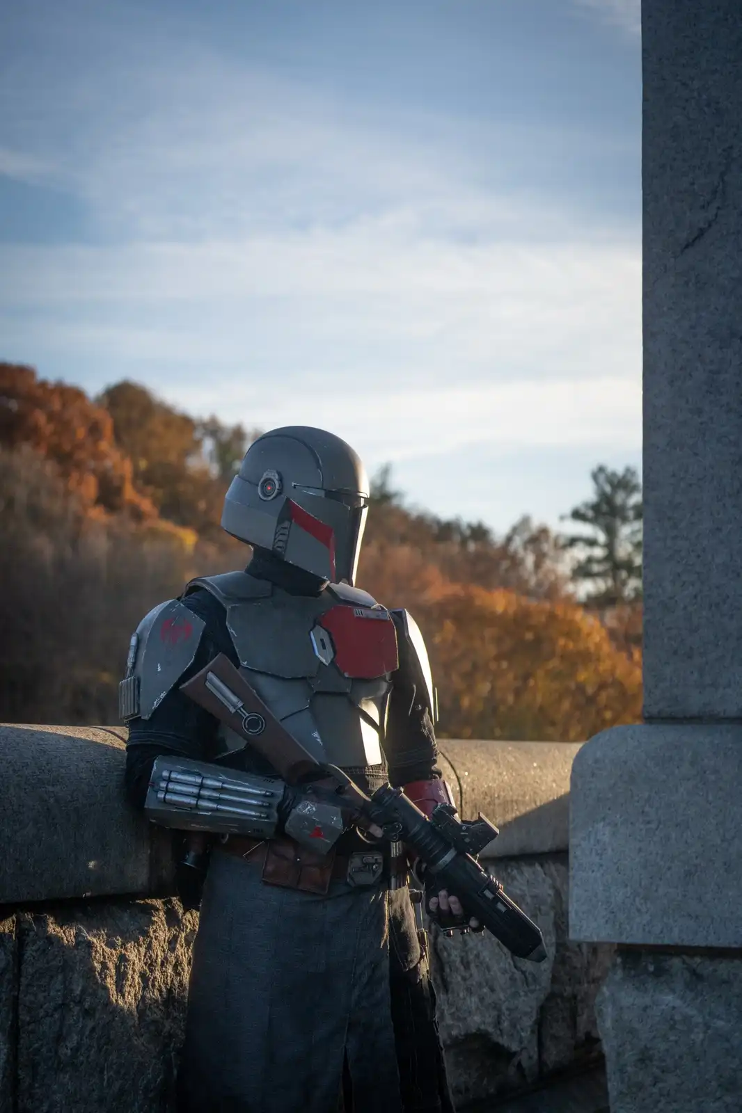 A person in detailed Mandalorian armor, featuring a helmet with a red stripe and a chest plate with red accents, stands…