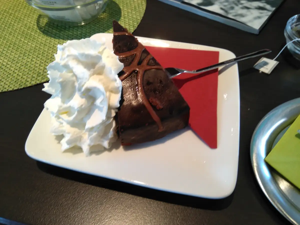 A slice of chocolate cake with chocolate drizzle and a large dollop of whipped cream sits on a white plate with a red…