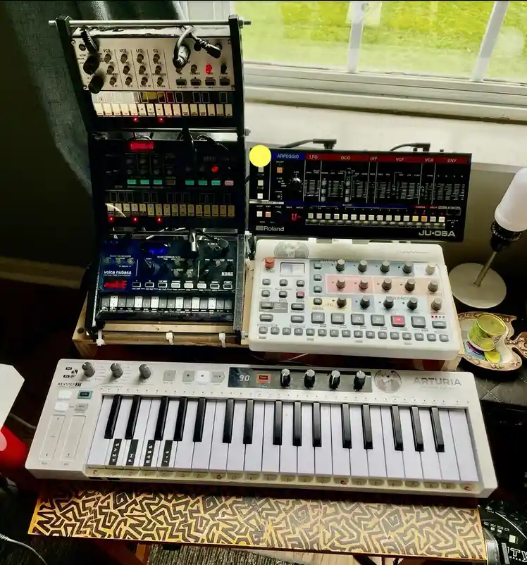 A photo shows a cluttered music production desk with multiple synthesizers and a keyboard. Visible gear includes a Korg…