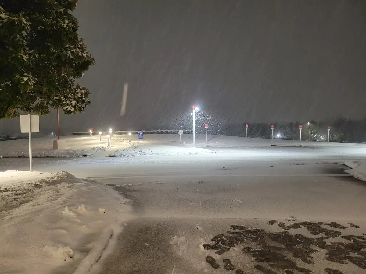 A nighttime photo of a snow-covered parking lot with heavy snowfall, illuminated by streetlights. Footprints are visible in…