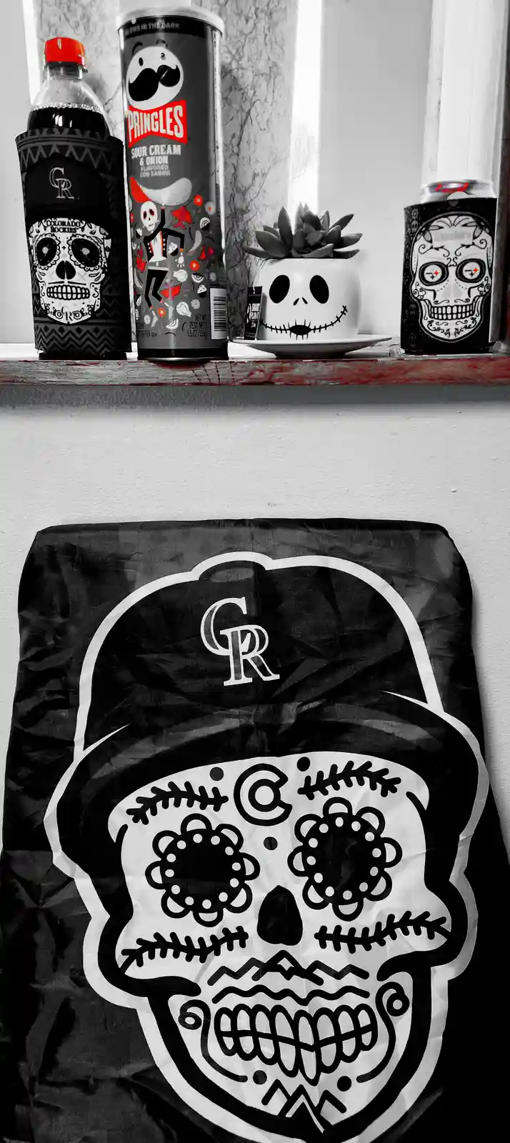 A black-and-white photo shows a shelf with a Pringles can, a soda bottle, and a beer can, all decorated with sugar skull…