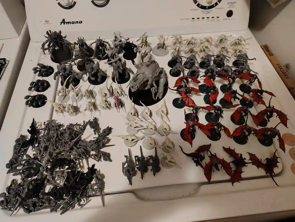 A white Amana washing machine top is covered with numerous unpainted and painted fantasy miniatures, arranged in sections…