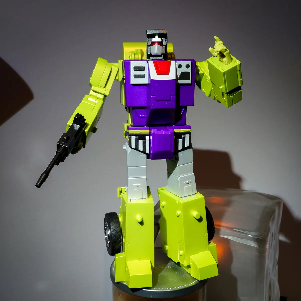 A lime green and purple transformer toy stands in robot mode, holding a black gun in one hand and making a peace sign with…