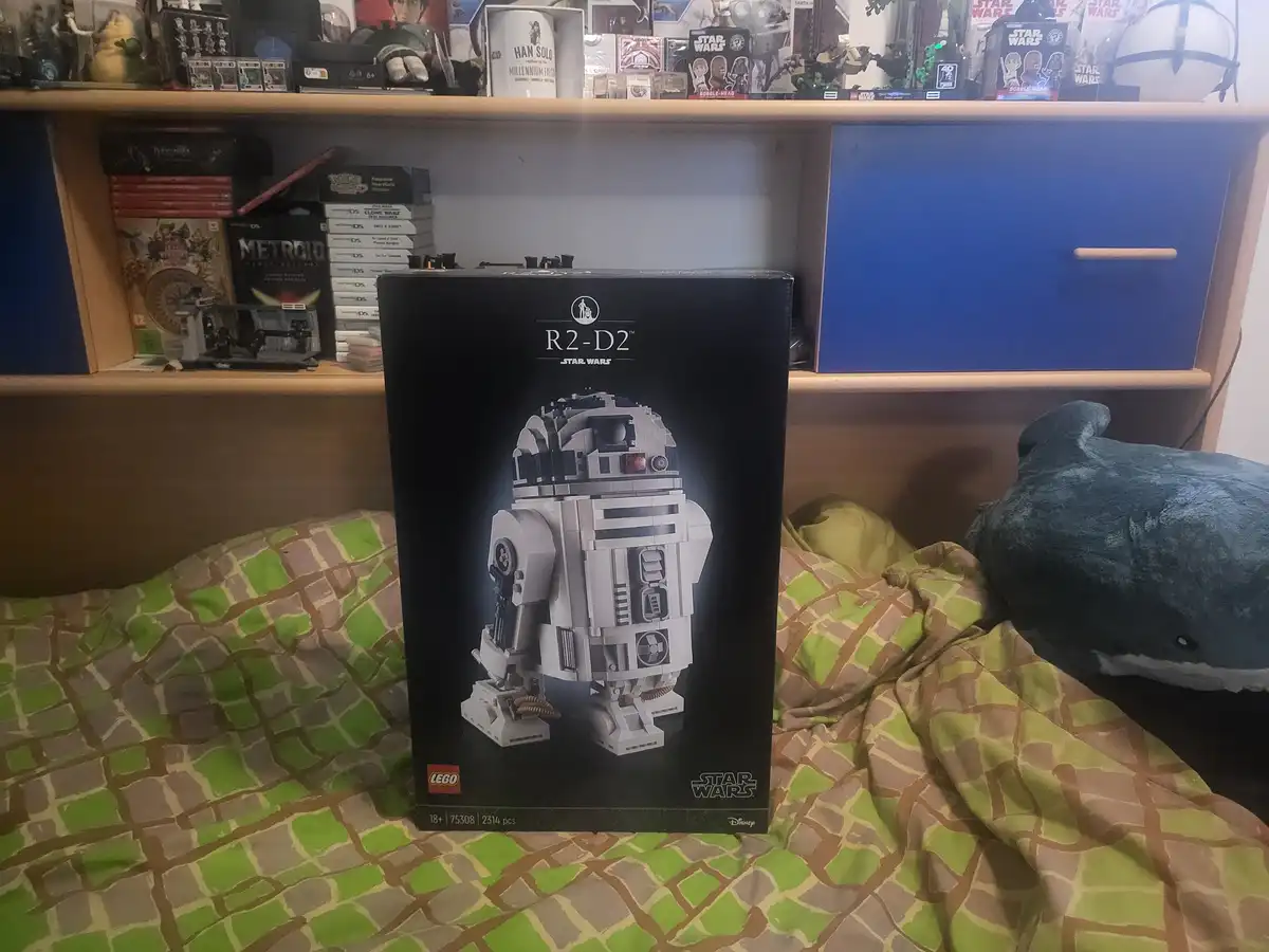 A large black LEGO Star Wars R2-D2 set box (set number 75308, 2314 pieces) stands upright on a bed with a green and brown…