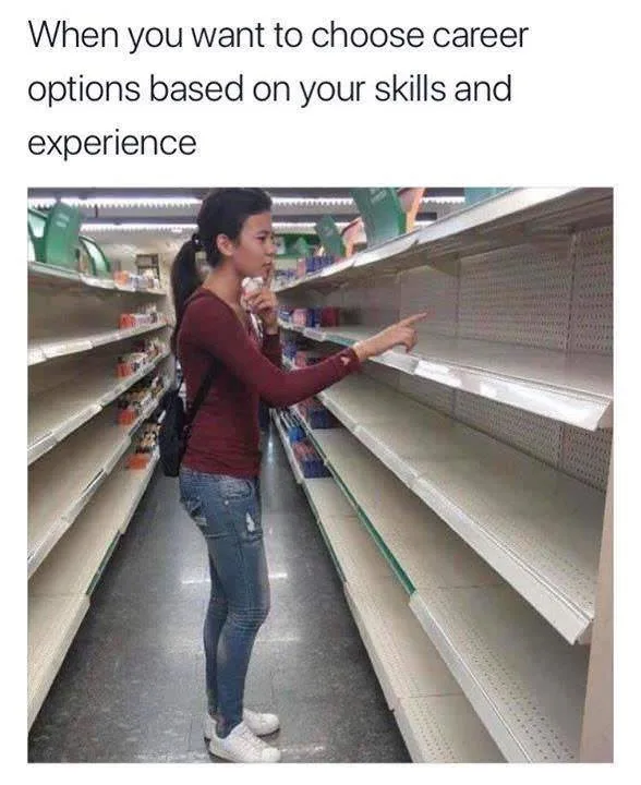 A woman stands in a supermarket aisle with mostly empty shelves, pointing at the bare shelves while holding a small item…