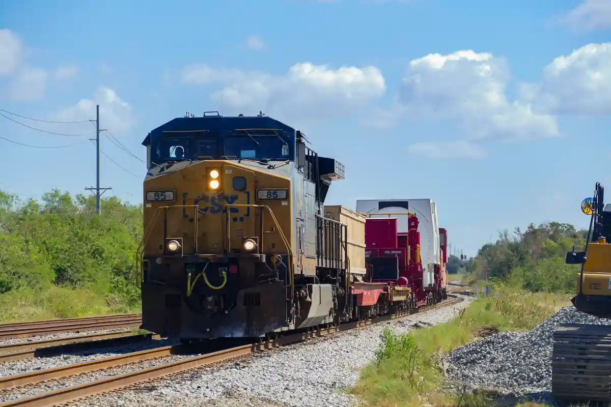 A CSY railroad locomotive numbered 85 is shown approaching on tracks under a partly cloudy sky, with a large white cargo…