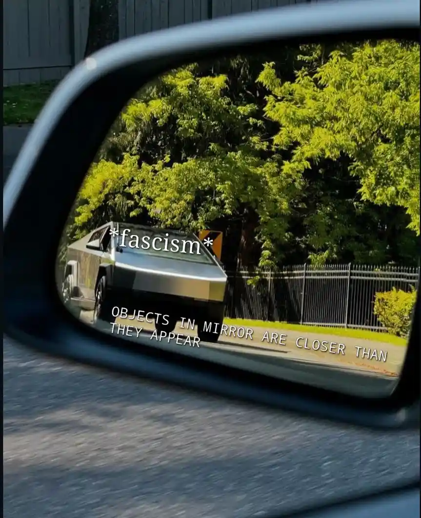 A car's side-view mirror reflects a silver Tesla Cybertruck parked behind a fence, with trees in the background. The…