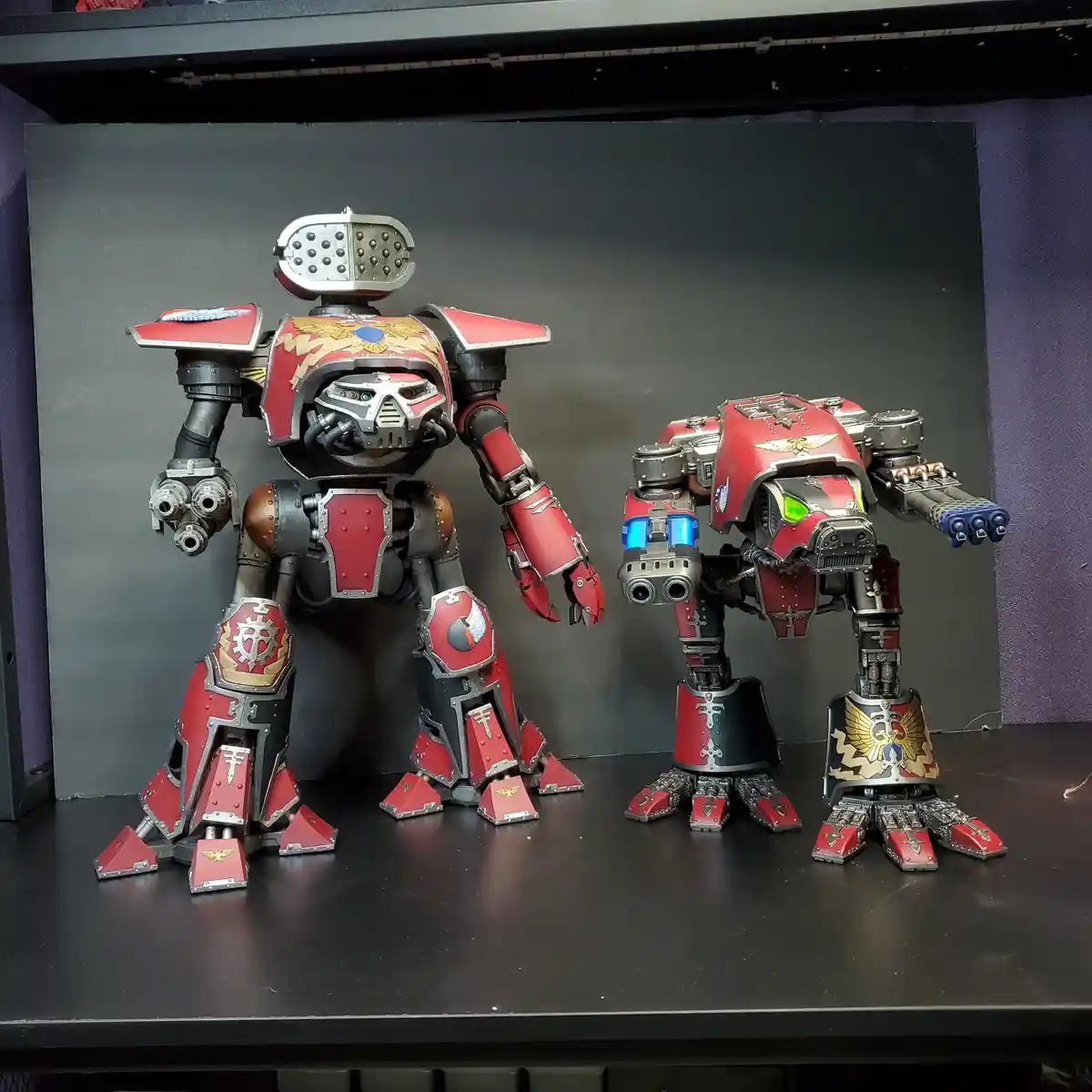 Two large, red and silver painted sci-fi mecha models stand side-by-side on a black shelf against a dark gray backdrop. The…