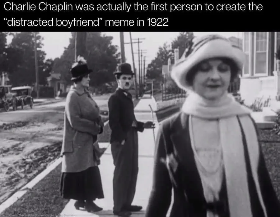 A black-and-white historical photo shows Charlie Chaplin in his iconic bowler hat and mustache, standing on a sidewalk and…