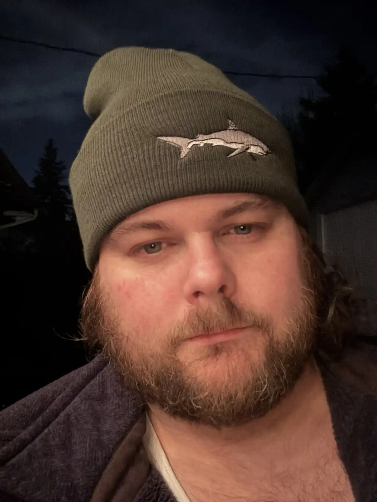 A man with a beard and long hair wears a green knit beanie featuring an embroidered shark logo. He looks directly at the…