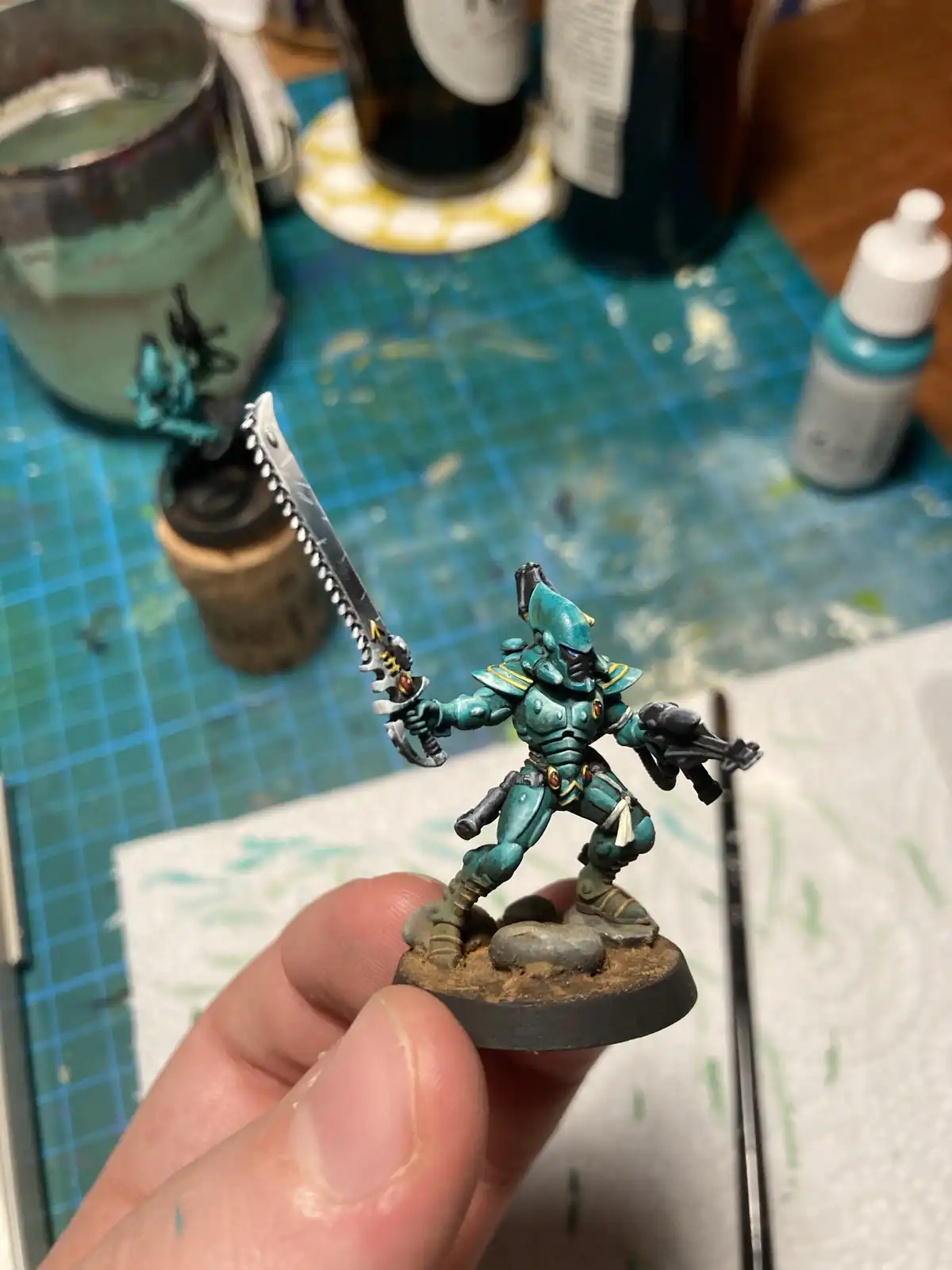 A hand holds up a freshly painted teal and gold fantasy miniature warrior wielding a serrated sword and a pistol, set…