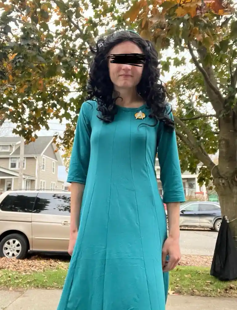 A person stands outdoors in a teal dress with a Starfleet insignia pin, their face obscured by black scribbles. The…