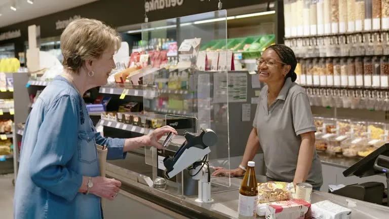 An older woman at a grocery store checkout is smiling and inserting her card into a modern card reader, while the cashier…