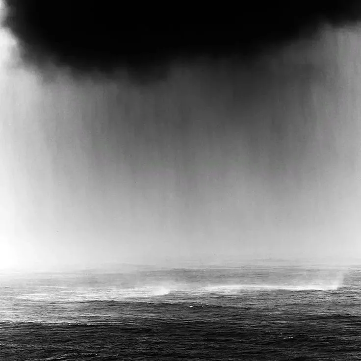 A black-and-white photograph shows a massive, dark storm cloud pouring rain directly onto a choppy ocean surface. The image…
