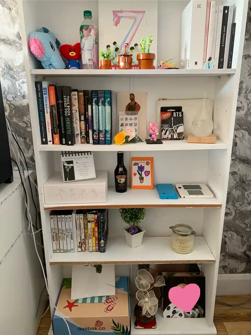 A white bookshelf filled with books, BTS merchandise, plush toys, a Baileys bottle, a Nintendo DS, a pink heart sticker, and…