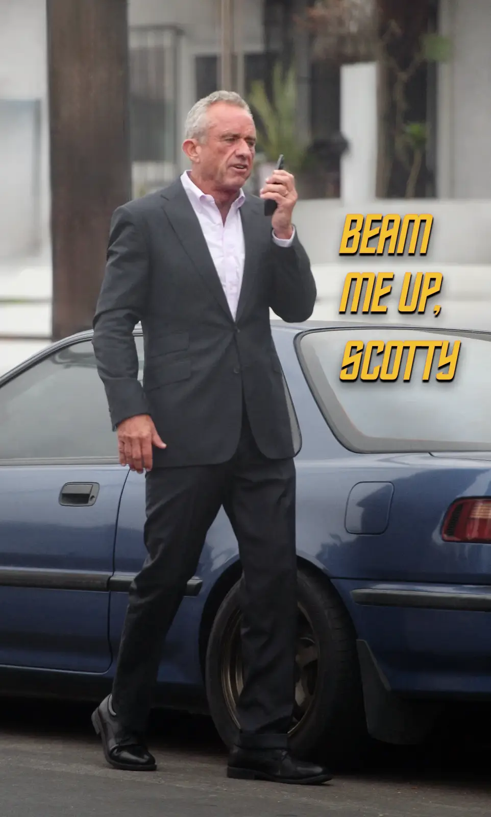 A man in a dark suit and light pink shirt stands beside a blue car, holding a phone to his ear as if calling for transport…