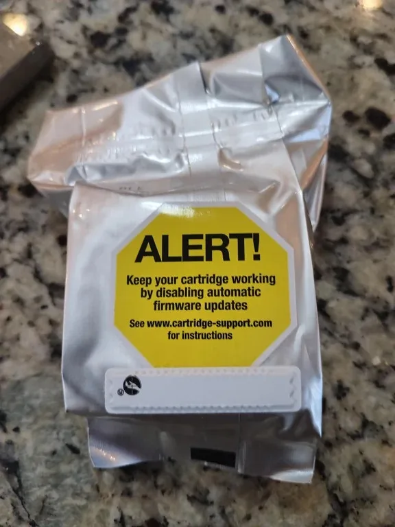 A silver-wrapped printer cartridge sits on a granite countertop, featuring a prominent yellow octagonal warning label. The…